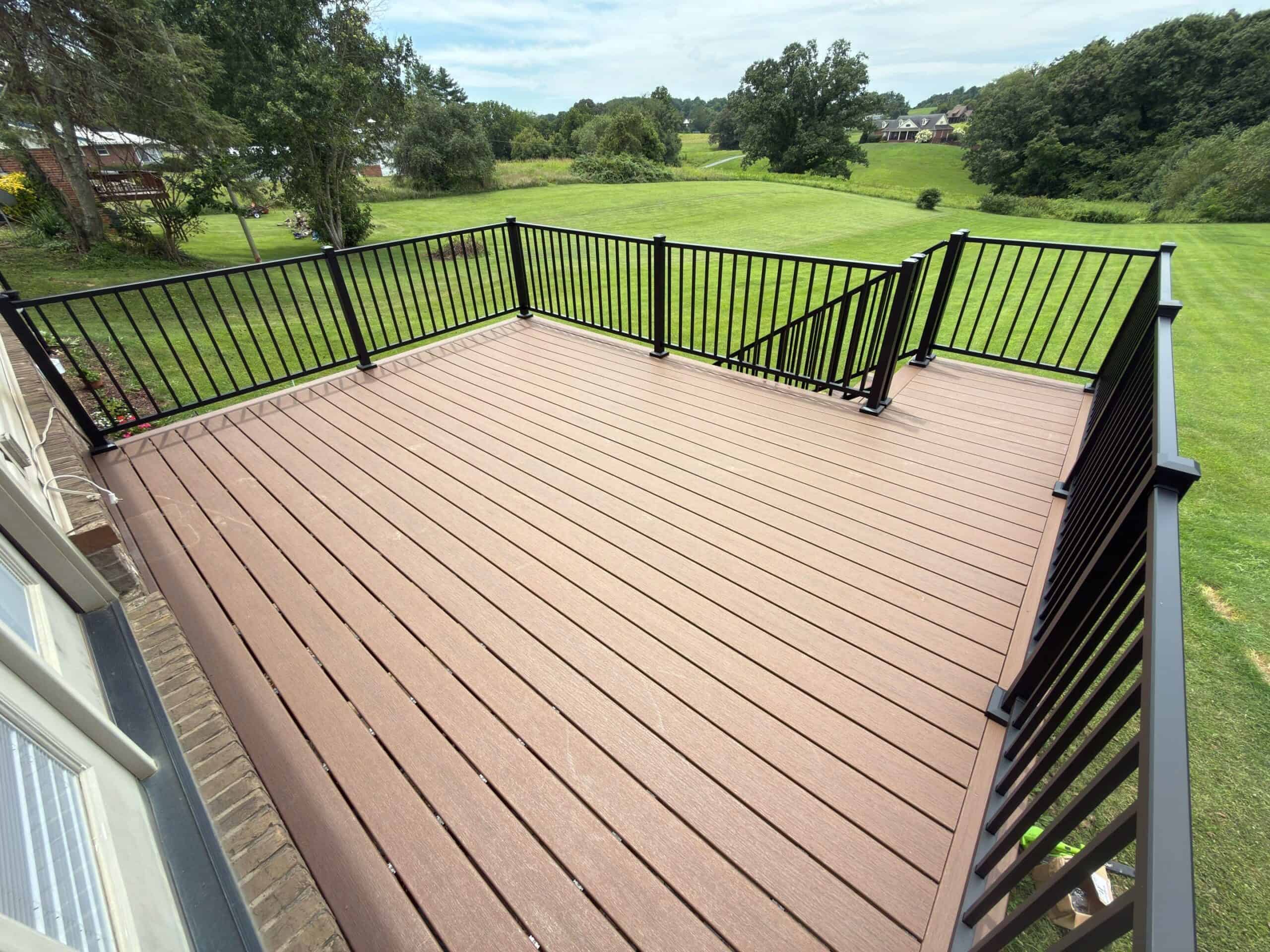 View from a second-story wooden deck with brown planks and black railing, overlooking a spacious green lawn and trees in the distance.